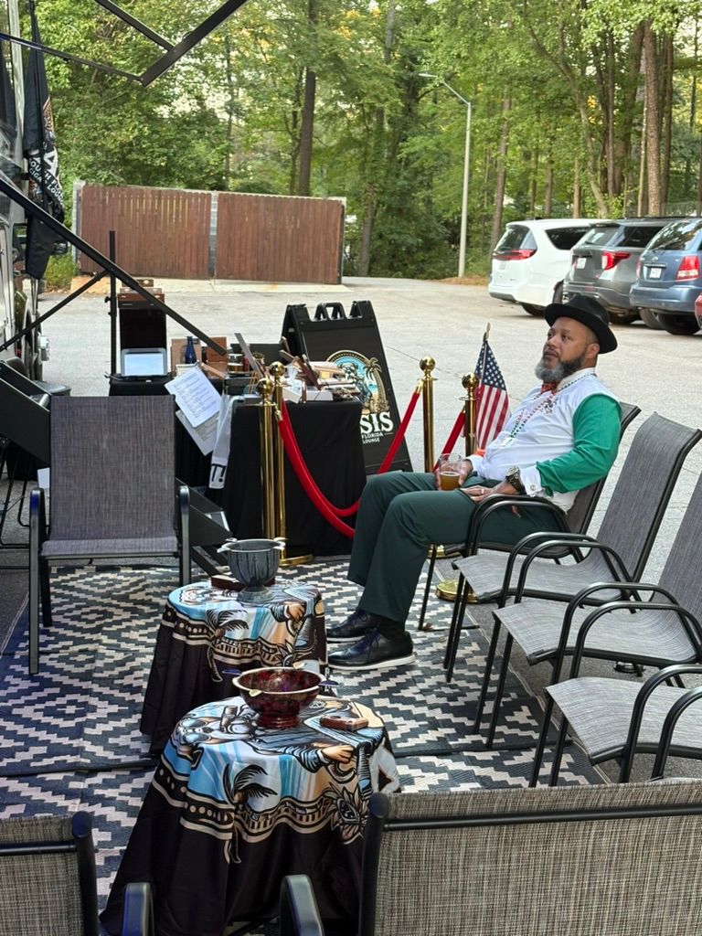 Outdoor parking-lot lounge under an RV awning: a man in a hat relaxes with a drink among patio chairs, patterned rugs, small draped tables, red ropes and American flags with a wooded backdrop.
