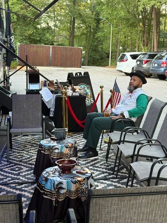 Outdoor parking-lot lounge under an RV awning: a man in a hat relaxes with a drink among patio chairs, patterned rugs, small draped tables, red ropes and American flags with a wooded backdrop.