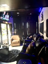 Cozy modern home theater lounge with black leather recliners, a beige armchair, wall-mounted TV showing sports commentary, blue accent lighting and a glass entry door