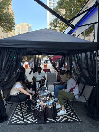 Five people relaxing under a black canopy tent on a downtown city street, seated around a patterned rug and low table with hookah and drinks at an outdoor street festival