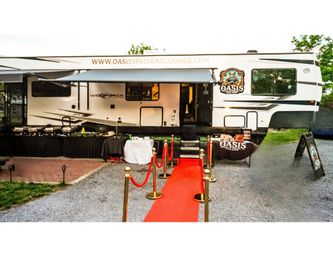 Mobile cigar lounge trailer at an outdoor pop-up event with awning, red carpet, gold stanchions and buffet tables on gravel