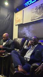 Two well-dressed people relaxing in a dim, upscale cigar lounge—leather recliners, blue neon glow, one exhaling cigar smoke, wall art of whiskey and a bulldog, small side table with a glass.