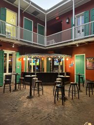 Cozy two-story courtyard bar at dusk with green shutters, white balcony, brick floor, string lights, patio heaters and high-top tables with stools.