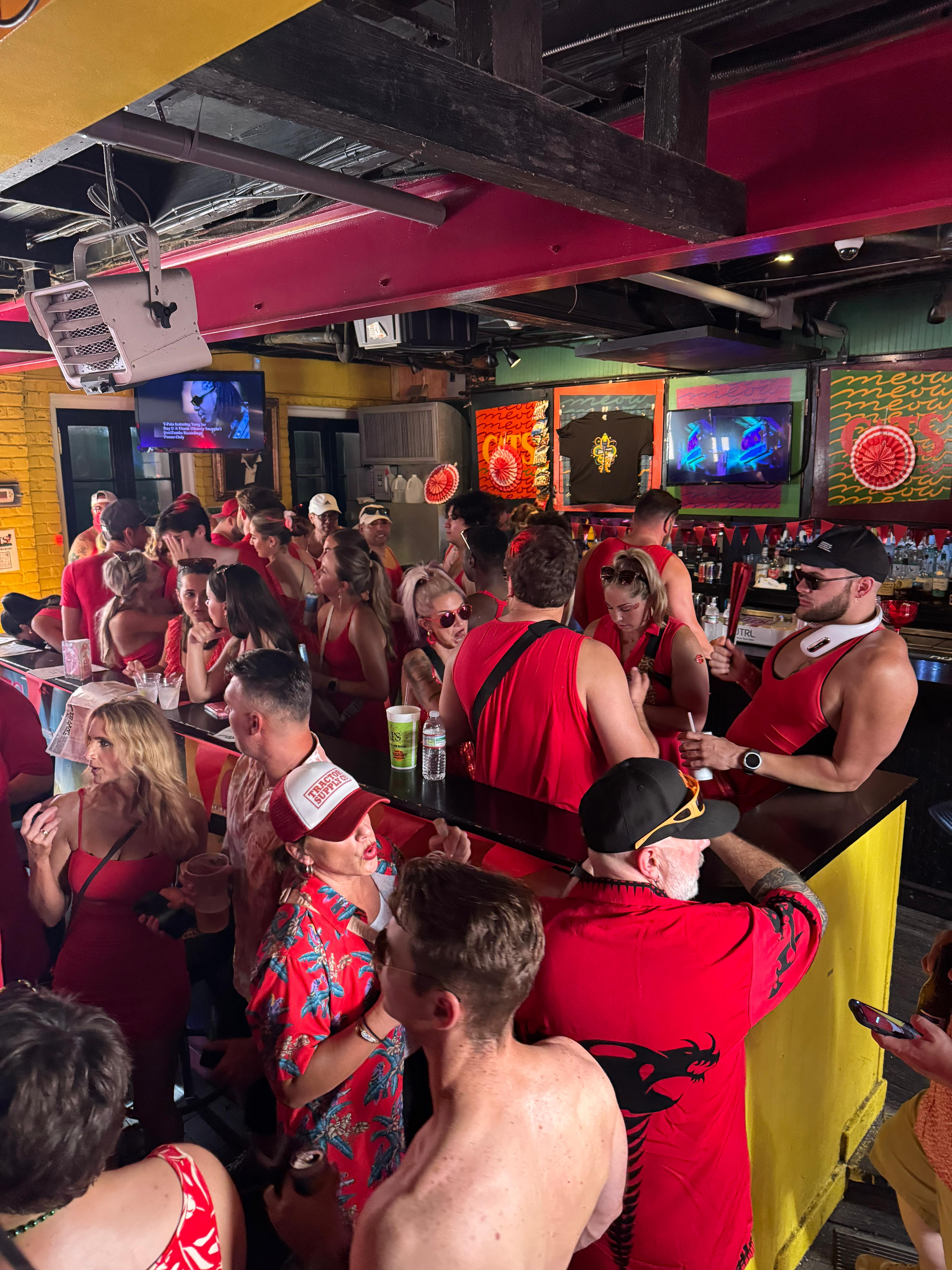 Packed indoor tropical‑themed bar with a lively crowd dressed mostly in red, people drinking and chatting at a busy counter beneath colorful artwork and TVs.