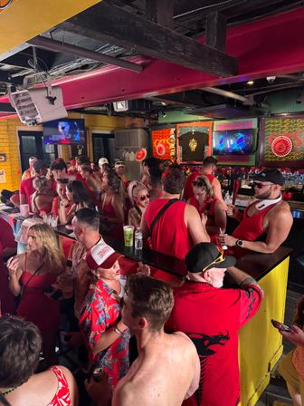 Packed indoor tropical‑themed bar with a lively crowd dressed mostly in red, people drinking and chatting at a busy counter beneath colorful artwork and TVs.