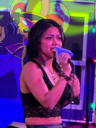Young woman singing into a microphone on a neon-lit karaoke stage with a colorful jester mural backdrop and string lights