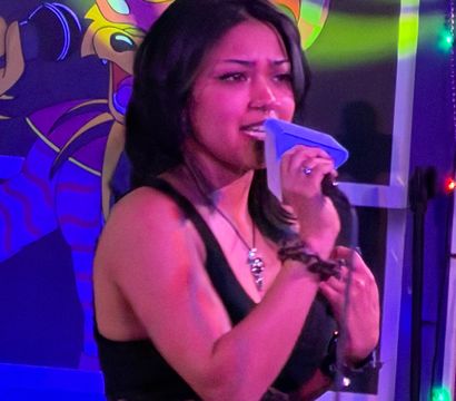 Young woman singing into a microphone on a neon-lit karaoke stage with a colorful jester mural backdrop and string lights
