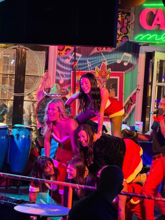 Women in Santa-style outfits forming a playful human pyramid on a colorful neon-lit bar stage with conga drums, music-note mural and a cheering crowd.