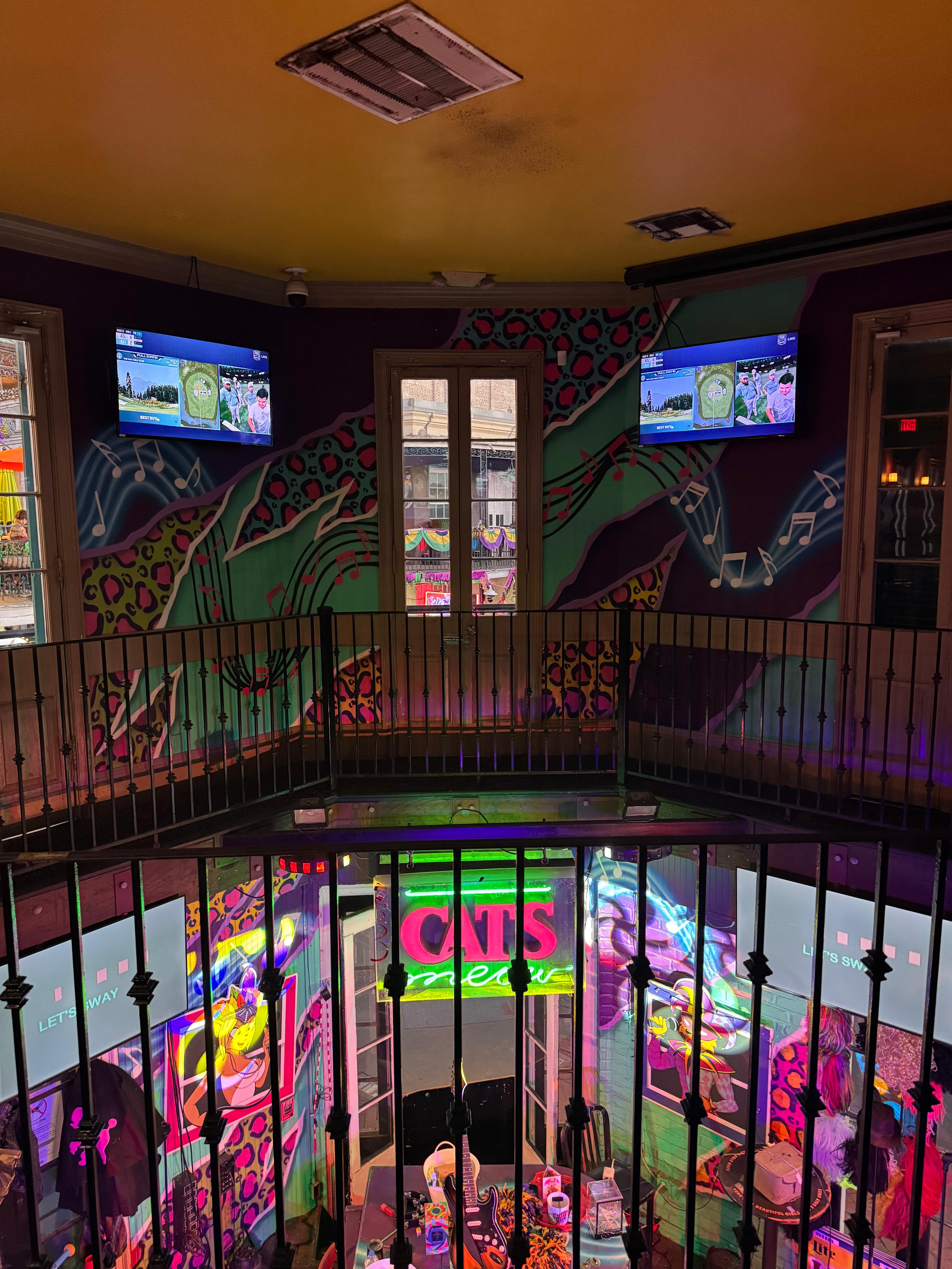 Colorful two-level music bar interior with wrought-iron balcony, neon pink-and-green sign over a small stage, leopard-print and musical-note wall mural, mounted TVs, and neon-lit guitars and decor below.