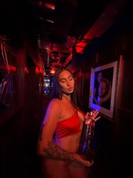 Woman in a red satin crop top puckering her lips while holding a tall bottle in a dim, red-lit nightclub corridor with framed portraits on the wall and visible arm tattoos, party vibe.