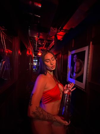 Woman in a red satin crop top puckering her lips while holding a tall bottle in a dim, red-lit nightclub corridor with framed portraits on the wall and visible arm tattoos, party vibe.