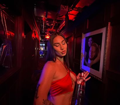 Woman in a red satin crop top puckering her lips while holding a tall bottle in a dim, red-lit nightclub corridor with framed portraits on the wall and visible arm tattoos, party vibe.