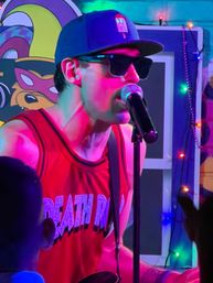 Performer in a baseball cap and sunglasses singing into a microphone on a small indoor stage, wearing a red graphic tank top amid colorful neon and string lights with audience silhouettes.