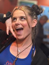 Playful close-up portrait of a young woman with space-buns and braids, septum and double-tongue piercings, bold winged eyeliner, wearing a blue tank top and layered necklaces, smiling animatedly in a lively indoor setting