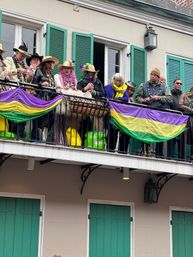 Group of people on a New Orleans balcony with green shutters, wearing festive hats, colorful beads and scarves as purple-green-gold Mardi Gras banners drape the wrought-iron railing.