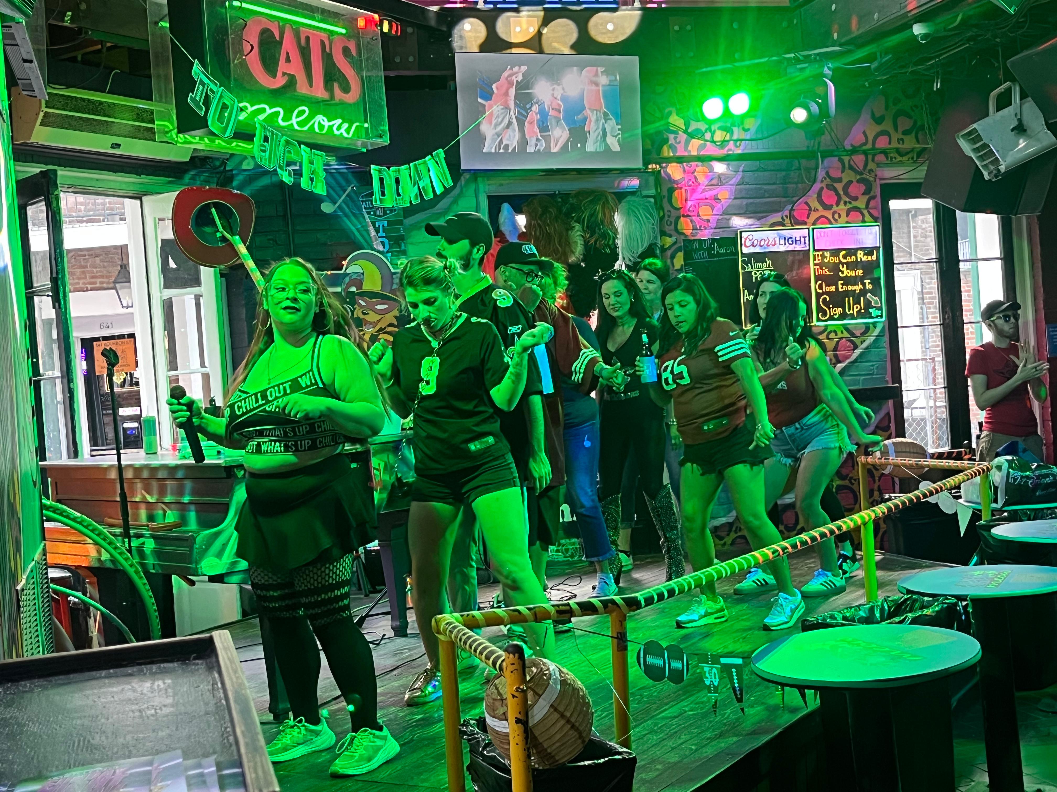 Karaoke night at a neon-lit downtown bar: a group of friends singing and dancing on a small stage under green party lights with microphones and cheering crowd.
