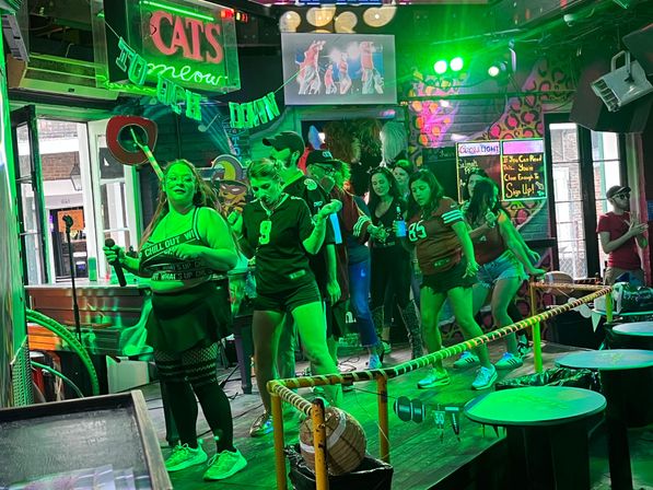 Karaoke night at a neon-lit downtown bar: a group of friends singing and dancing on a small stage under green party lights with microphones and cheering crowd.