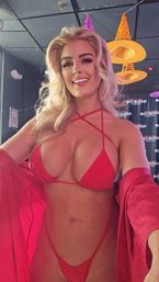 Blonde woman smiling selfie in a red strappy bikini and sheer robe inside a nightclub with hanging witch-hat decorations, Halloween-themed party vibe.