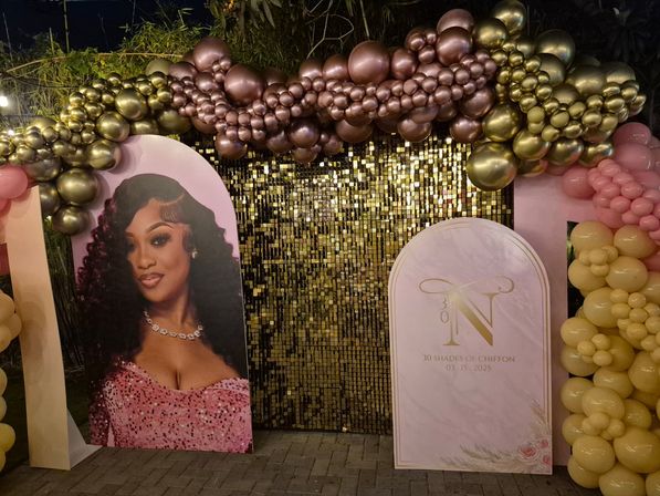 Outdoor party backdrop with shimmering gold sequin wall framed by metallic rose-gold and gold balloon garlands, large photo cutout of a woman in a pink sequin dress, arched sign with gold monogram “N” and text “30 Shades of Chiffon 03.15.2025,” and clusters of pink and yellow balloons.