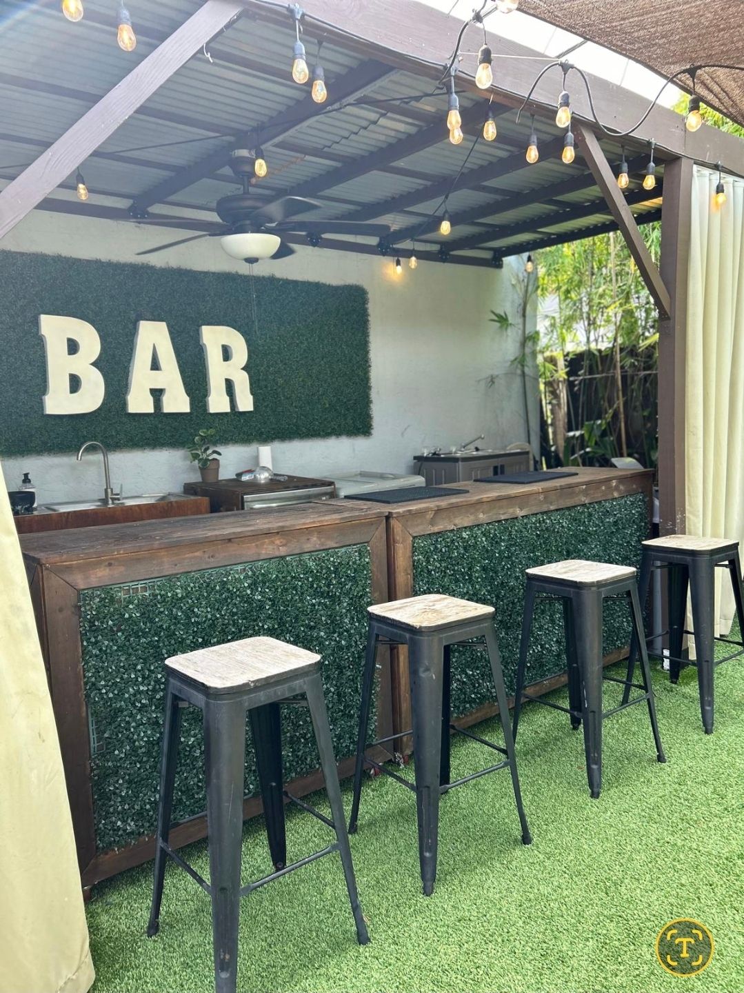 Cozy backyard patio bar under a pergola with Edison string lights, wooden counter wrapped in faux boxwood, four metal bar stools on green artificial turf, ceiling fan and large “BAR” letters on the wall.