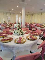 Chic indoor wedding reception in a banquet hall with round tables draped in white linen, pink chair covers and napkins, gold charger plates and flatware, tall glass candle holders and white-and-pink floral centerpieces, fairy lights and hanging greenery on sheer curtains.