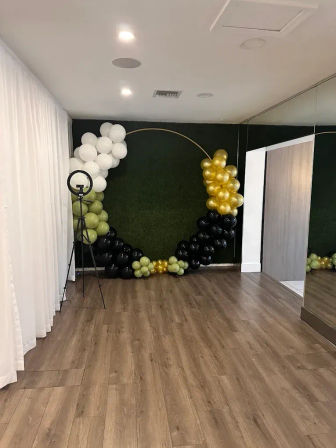 Chic indoor party photo backdrop with a gold circular hoop and white, gold, black, and sage-green balloon arch against a faux hedge wall, wood floor and ring light
