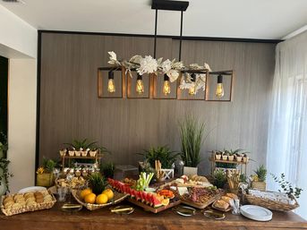 Mouthwatering rustic indoor buffet on a wooden table beneath a floral Edison-bulb chandelier, featuring charcuterie boards with cheeses, cured meats, crackers and bread, sliced watermelon and citrus, vegetable sticks and dips, mini desserts, potted greenery and plates by a sheer-windowed dining area.