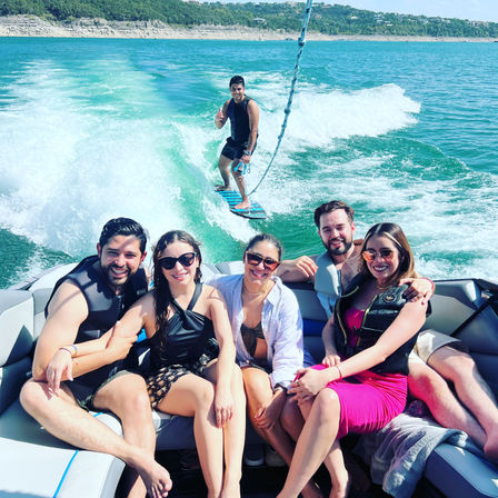 Smiling group of friends lounging on a speedboat while a wakesurfer rides the turquoise wake on a sunny summer lake with a tree-lined shoreline.