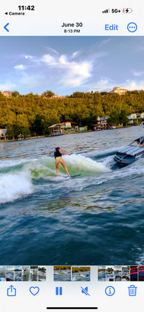 Person wake-surfing behind a speedboat on a lake at sunset, riding a board through a greenish wake with tree-covered shoreline and lakeside homes in the background.