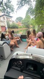 Group of friends in swimsuits on a pontoon boat cruising a tree-lined residential canal past Mediterranean-style homes on a sunny summer day.