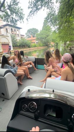 Group of friends in swimsuits on a pontoon boat cruising a tree-lined residential canal past Mediterranean-style homes on a sunny summer day.
