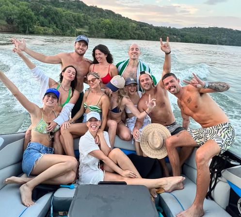 Group of friends in swimsuits posing and laughing on a speedboat cruising a tree-lined lake at sunset with a wake trailing behind — fun summer boat party vibe.