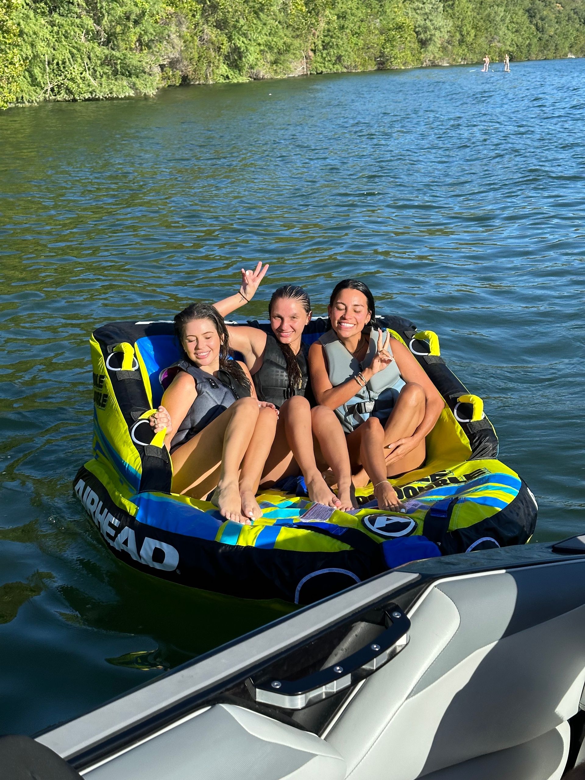Three friends in life jackets smiling and flashing peace signs while sitting on a colorful inflatable towable behind a boat on a sunny lake with a tree-lined shoreline.