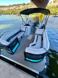 Pontoon boat docked under a bridge on a sunny lake, gray wraparound seating and black canopy with teal-lit accents, tree-lined cliffs and other boats in the background.