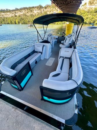 Pontoon boat docked under a bridge on a sunny lake, gray wraparound seating and black canopy with teal-lit accents, tree-lined cliffs and other boats in the background.