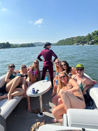 Eight friends on a pontoon boat enjoying summer drinks on a sunny lake with wooded shoreline and hills, one person in a burgundy suit striking a pose at the bow
