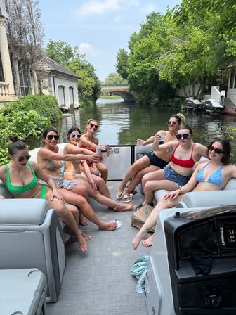 Seven friends in swimsuits toasting drinks on a pontoon boat cruising a tree-lined canal past riverfront homes and a stone bridge on a sunny summer day