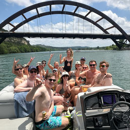 Pontoon boat party with a smiling group in swimsuits holding drinks under a large arched bridge on a sunny summer day over a calm river with green hills in the background.