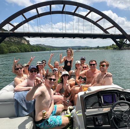 Pontoon boat party with a smiling group in swimsuits holding drinks under a large arched bridge on a sunny summer day over a calm river with green hills in the background.
