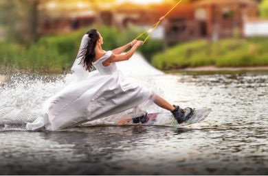 Bride wakeboarding across a lake in a flowing white wedding dress and veil, gripping a tow rope as water sprays behind her — playful outdoor water sports moment.