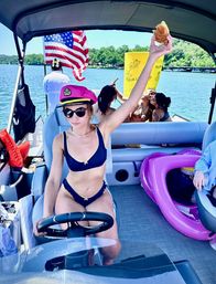 Summer lake day on a pontoon boat — woman in a pink captain's hat and navy bikini steers while holding up a sandwich, with an American flag, inflatable floats and friends lounging in the background.