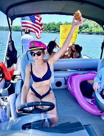 Summer lake day on a pontoon boat — woman in a pink captain's hat and navy bikini steers while holding up a sandwich, with an American flag, inflatable floats and friends lounging in the background.