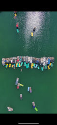 Aerial drone view of a summer lake raft-up: a long line of colorful pontoons and small boats tied together with bright floating mats and swimmers on green water, sunlight sparkling on the surface.