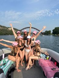 Seven women in bikinis wearing matching pink baseball caps celebrate with arms raised aboard a pontoon boat on a sunny lake, with an arched bridge and tree-lined shoreline in the background.