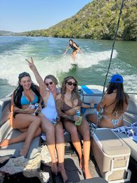 Four friends in swimsuits relaxing on a motorboat while a person wakesurfs on the boat’s wake on a sunny lake with tree-covered hills in the background.