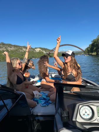 Group of friends in swimsuits partying on a motorboat, raising hands under a clear blue sky with an arched bridge and green shoreline in the background on a sunny lake day
