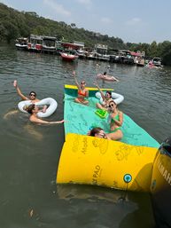Group of friends at a summer lake party lounging on colorful floating mats and inflatables, sipping drinks and waving near a row of anchored pontoon boats under sunny skies.