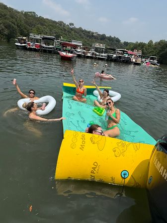 Group of friends at a summer lake party lounging on colorful floating mats and inflatables, sipping drinks and waving near a row of anchored pontoon boats under sunny skies.