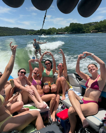 Group of friends in bikinis on a speedboat enjoying a sunny lake day—cheering and making heart-hand shapes while a wakesurfer rides the boat wake with tree-lined shore in the background.