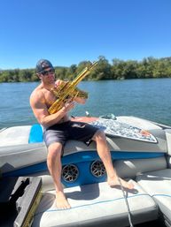 Shirtless man wearing sunglasses and a backwards cap sitting on a boat at a sunny lake, holding a shiny gold inflatable toy rifle beside a wakeboard under a clear blue sky.